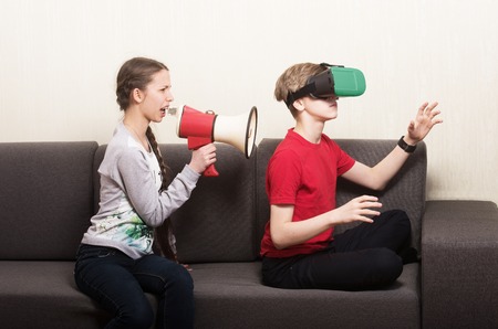 Young girl shouting through the megaphone at the boy wearing virtual reality 3D glasses, sitting on the sofa. Ignore real communication concept.の写真素材