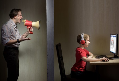 Tween son in headphones looks at a computer screen while his father yells at him through a megaphoneの写真素材