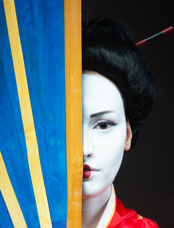 Woman in geisha makeup covering half of her face with a big fanの写真素材