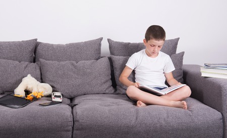 Boy reads an interesting book of the sofa. Studio portrait on white background.の写真素材