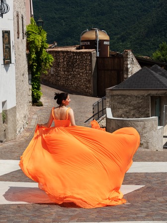 Beautiful girl in amazing fluttering orange dress running forward in old town.の写真素材