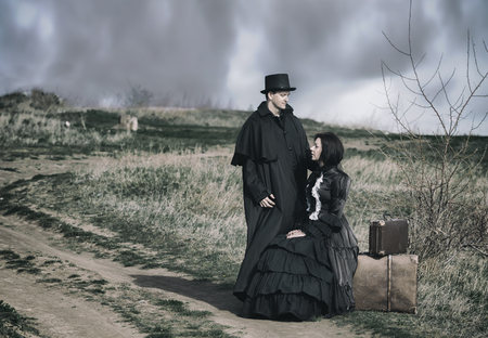 Outdoors portrait of a victorian lady in black sitting on the road with her luggage and gentleman...