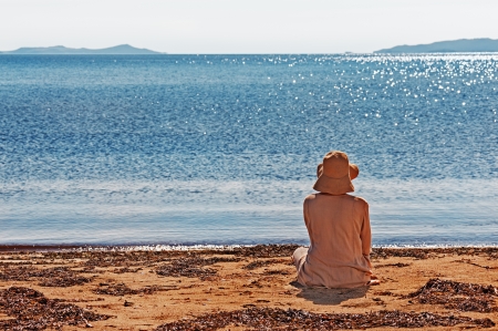 Portrait of young woman on the beach near the sea sitting wearing dress and hat. Photo from behind.の写真素材