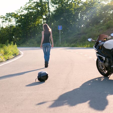 Young beautiful blonde girl in trendy jeans and a black T-shirt took off and put his helmet on the road near the modern motorcycle. Outdoor portrait in soft sunlight.の写真素材
