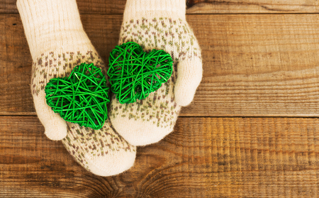 Woman hands in light teal knitted mittens are holding green hearts on wooden background. Winter and Christmas concept.の写真素材