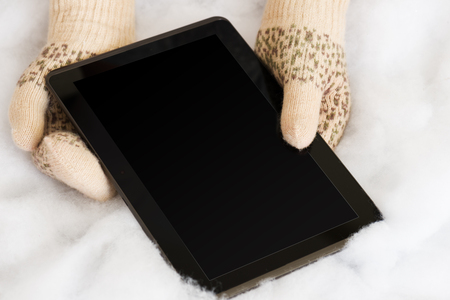 Woman hands in light teal knitted mittens are holding modern tablet pc on snow background. Winter concept.の写真素材