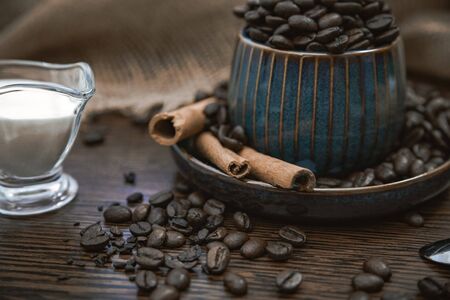 a blue ceramic cup full of coffee beans with cinnamon sticks, scattered coffee beans, milk and canvas fabric on brown wooden tableの写真素材