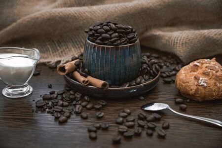 a blue ceramic cup full of coffee beans, with cinnamon sticks and spoon, scattered coffee beans, bun, milk and canvas fabric on brown wooden tableの写真素材