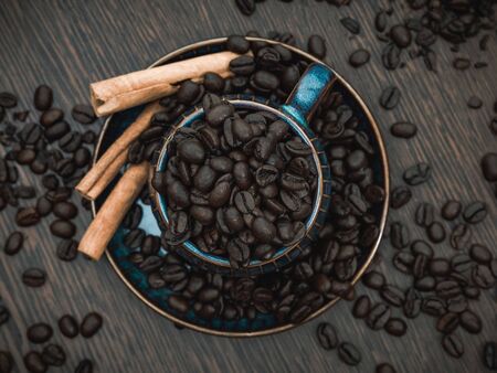 a blue ceramic cup full of coffee beans with cinnamon sticks and scattered coffee beans on brown wooden tableの写真素材