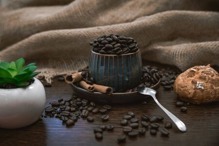 a blue ceramic cup full of coffee beans, with cinnamon sticks and spoon, scattered coffee beans, plant, cream bun and canvas fabric on brown wooden tableの写真素材