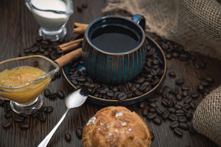 a blue ceramic cup with americano, cinnamon sticks, orange jam, spoon, scattered coffee beans, bun, milk and canvas fabric on brown wooden tableの写真素材