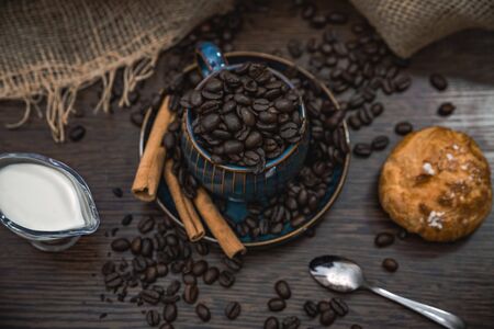 a blue ceramic cup full of coffee beans with cinnamon sticks, spoon, scattered coffee beans, bun, milk and canvas fabric on brown wooden tableの写真素材