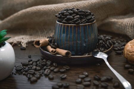 a blue ceramic cup full of coffee beans, with cinnamon sticks and spoon, scattered coffee beans, plant, bun and canvas fabric on brown wooden tableの写真素材