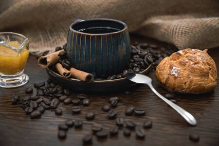 a blue ceramic cup with americano, cinnamon sticks, orange jam, spoon, scattered coffee beans, bun and canvas fabric on brown wooden tableの写真素材
