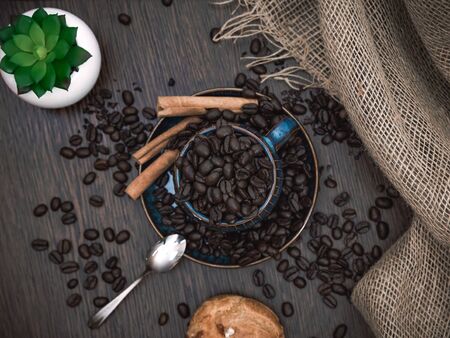 a blue ceramic cup with coffee beans, cinnamon sticks and spoon, scattered coffee beans, plant, bun and canvas fabric on brown wooden tableの写真素材