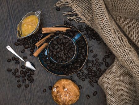a blue ceramic cup full of coffee beans with cinnamon sticks, orange jam, spoon, scattered coffee beans, bun and canvas fabric on brown wooden tableの写真素材