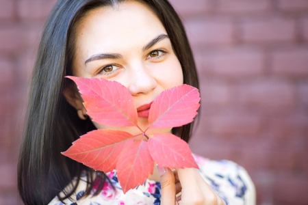Pretty Girl hiding behind red autumn leaf a brick wall background. Autumn Concept