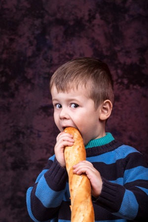 Cute kid holding and biting French bread against dark red background. Boy with appetite eats bread. Little boy eating the french baguetteの写真素材