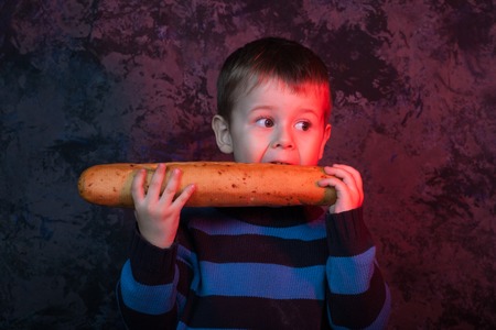 Cute kid holding and biting French bread against dark red background. Boy with appetite eats bread. Little boy eating the french baguetteの写真素材