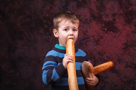 Cute kid holding and biting French bread against dark red background. Boy with appetite eats bread. Little boy eating the french baguetteの写真素材