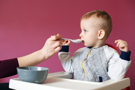 Mom feeds her beautiful baby with a spoon. Caring good mother feeds her little child son with a...