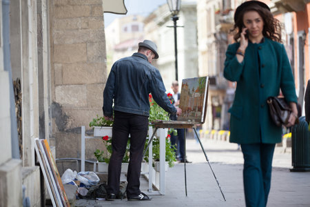 Lviv, Ukraine - April, 2016: Artist paints a cityscape in the center Lviv. A beautiful woman talking on her phone and walking on the street in a European city. A man paints with oil on canvas.のeditorial素材