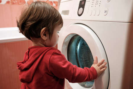 Baby girl peeking into washing machine at home. Little Caucasian baby girl 1 year with washing machine. Home life childhood.の写真素材