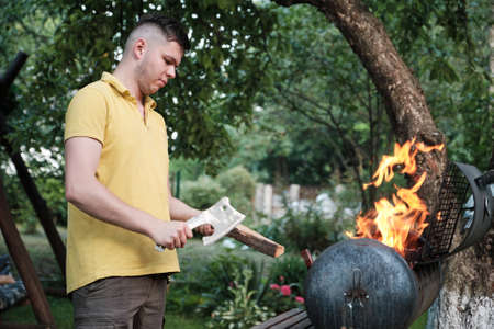 Young man putting firewood in mangal. Handsome athlete male builds a fire in a metal barbecue in the...