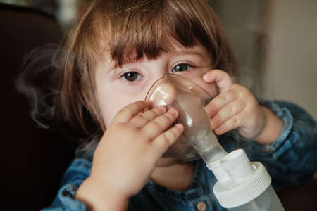 Little girl in a mask, treatments respiratory tract with a nebulizer at home. Baby sits with a nebulizer in his mouth, inhaler, treatment of bronchitis. Nebulizer, cute baby girl using inhaler.の写真素材