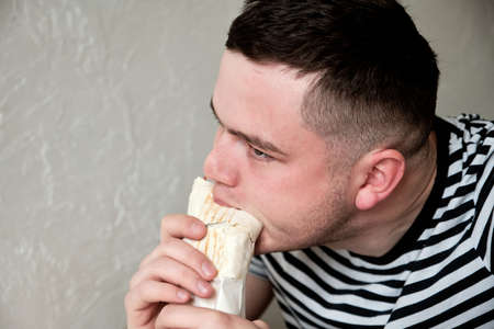 Handsome Young Man Eating Kebab. Hungry Man eating fast food indoors. Portrait of an attractive young man in kitchen wearing casual clothes.の写真素材