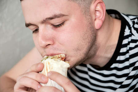 Handsome Young Man Eating Kebab. Hungry Man eating fast food indoors. Portrait of an attractive young man in kitchen wearing casual clothes.の写真素材