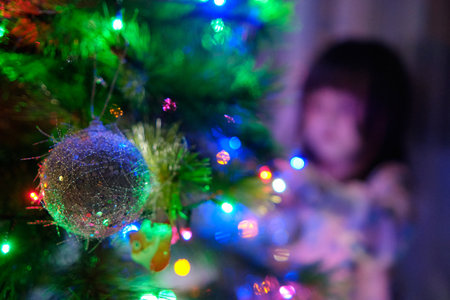 A 4-year-old charming little girl decorates a Christmas tree in light of garlands, her eyes sparkling with wonder as she hangs the ornaments. Cute baby girl hangs Toys on the Christmas Treeの写真素材
