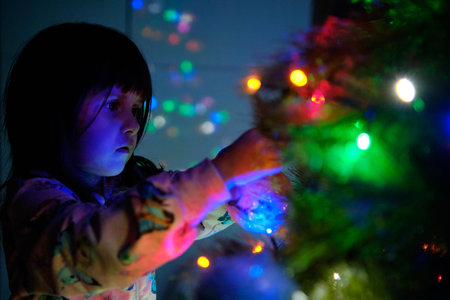 A 4-year-old charming little girl decorates a Christmas tree in light of garlands, her eyes...