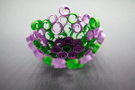 A close-up of a decorative bowl crafted from intricately rolled purple and green paper strips,...