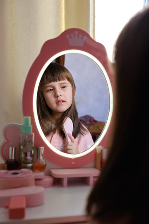 Little girl brushing her hair looking in a toy mirror with LED lights. Cute preschooler playing with vanity table and combing hair at home. Child's reflection in a pink toy mirror while grooming and playing pretendの写真素材