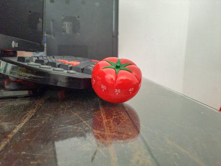 Pomodoro kitchen timer in form of a red tomato laying on a wooden table , with colorful objects...