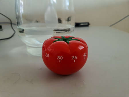 Pomodoro kitchen timer in form of a red tomato laying on a wooden table , with colorful objects aroundの写真素材