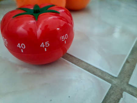 Pomodoro kitchen timer in form of a red tomato laying on a wooden table , with colorful objects aroundの写真素材