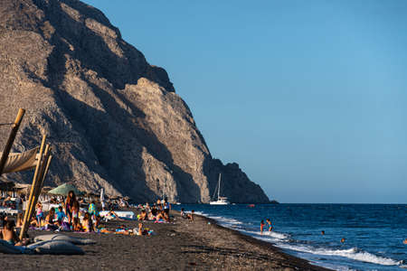 Perissa, Greece - July 11 2019:   Sun bathers and swimmers throng on the black volcanic sand of Perissa Beachのeditorial素材
