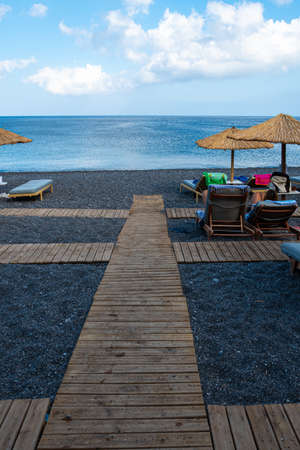 Kamari, Greece - July 14 2019:   A board walk leads across the black sand between the parasols and sunbeds on Kamari beachのeditorial素材