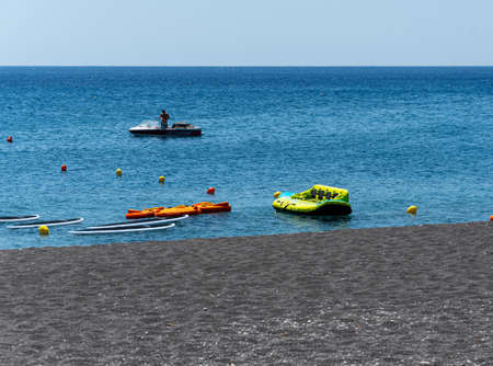 Perissa, Greece - July 18 2019:   An inflatable, some canoes and some surfboards awaiting holidaymakers with a speedboat behind them in the Aegean Sea at Perissaのeditorial素材