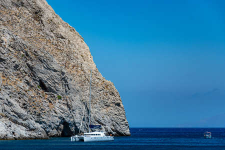 Perissa, Greece - July 11 2019:   A Yacht lies anchored in the Aegean sea beneath the cliffs at the...