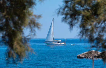 Perissa, Greece - July 13 2019:   A yacht in the blue waters of the Aegean Sea, seen between two tree on the Perissa Sea Front roadのeditorial素材