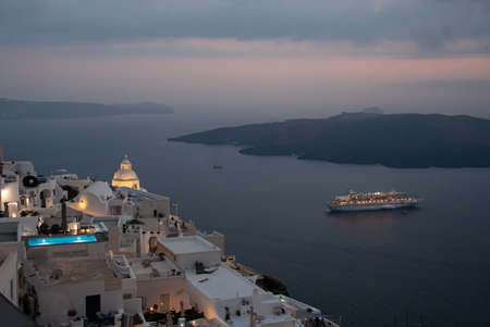 Fira, Greece - July 14 2019:   The twilight view over Fira's hotels and churches to the cruise liner moored in the Aegean Sea below, taken from Ipapantisのeditorial素材