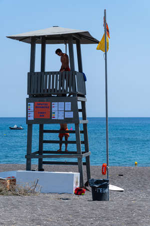 Perissa, Greece - July 11 2019:   Two life guards on duty on the tower on the black sand beach of...
