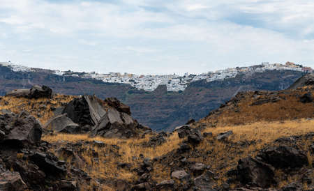 The black basalt rocks of Nea Kameni Island, the dome of the Santorini volcano with the towns of Santorini in the distanceの写真素材