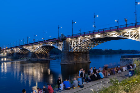 Warsaw, Poland - July 20, 2019: Group of people relax at the Vistula River waterfront by the Poniatowski Bridge (Polish: Most Poniatowskiego) illuminated at nightのeditorial素材