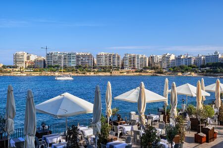 Outdoor restaurant tables in St. Julian town seaside terrace in Malta with view to Sliema town skyline across the sea bay.の写真素材