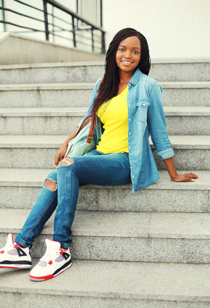 Beautiful happy smiling african woman wearing a jeans shirt and backpack resting on stairsの写真素材