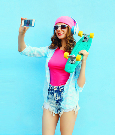 Fashion pretty cool girl with skateboard makes self portrait on smartphone over blue backgroundの写真素材